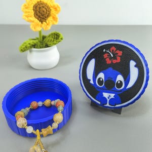 K&ouml;nnte beinhalten: Ein blauer Beh&auml;lter mit einem Perlenarmband und einem Untersetzer im Stitch-Design. Das Armband hat verschiedenfarbige Perlen und einen goldfarbenen Anh&auml;nger. Eine geh&auml;kelte Sonnenblume in einem wei&szlig;en Topf ist ebenfalls vorhanden.