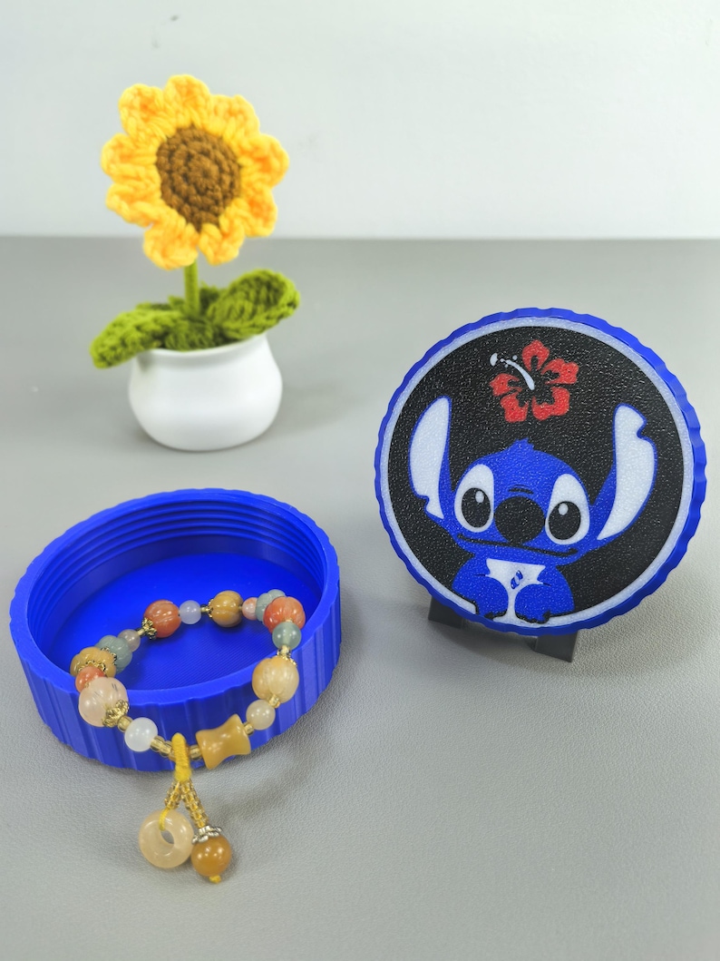 K&ouml;nnte beinhalten: Ein blauer Beh&auml;lter mit einem Perlenarmband und einem Untersetzer im Stitch-Design. Das Armband hat verschiedenfarbige Perlen und einen goldfarbenen Anh&auml;nger. Eine geh&auml;kelte Sonnenblume in einem wei&szlig;en Topf ist ebenfalls vorhanden.