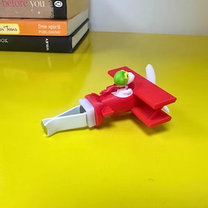 May include: A red and white toy airplane with a green pilot figure. The toy has a propeller and wings. The airplane is on a yellow surface. The toy is approximately 6 inches long.