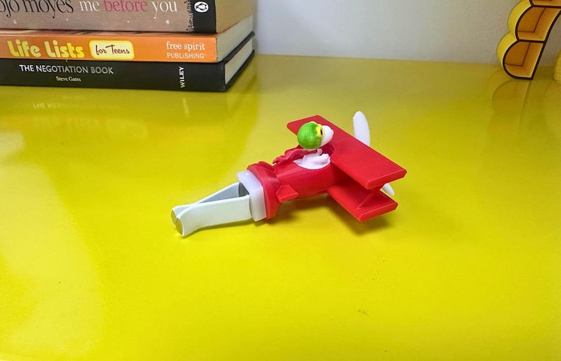 May include: A red and white toy airplane with a green pilot figure. The toy has a propeller and wings. The airplane is on a yellow surface. The toy is approximately 6 inches long.