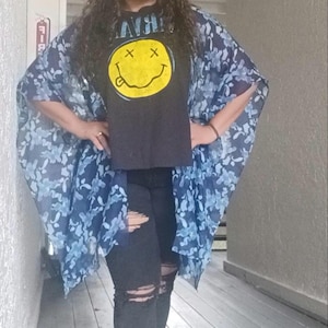 May include: A black t-shirt featuring a yellow smiley face graphic with X-shaped eyes and the text NIRVANA, layered under a blue and white camouflage-patterned sheer kimono-style cover-up, paired with black distressed denim jeans with knee rips.
