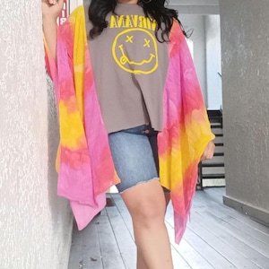 May include: A grey t-shirt featuring a yellow Nirvana smiley face graphic, paired with blue denim shorts and a vibrant pink, orange, and yellow tie-dye kimono. The outfit is completed with blue thong sandals and silver hoop earrings.
