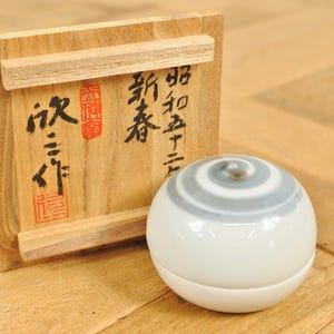 以下が含まれることがあります： 白いセラミック製の球体で、上部に灰色の渦巻き模様があり、日本の書道と赤いスタンプが付いた木箱の隣にあります。箱は明るい色の木で作られています。