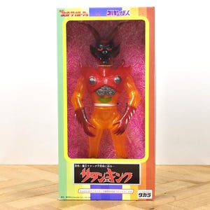 May include: A vintage action figure of a red and orange monster with a black head and horns, packaged in a colourful box. The box features Japanese text and a rainbow stripe design. The figure is posed inside the box.