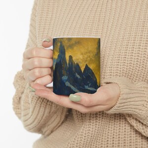 May include: White ceramic mug featuring a mountain landscape painting in shades of blue and gold. The artwork depicts jagged peaks against a golden sky. The mug is held in a person's hands.