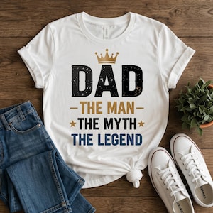 May include: A white short-sleeved t-shirt featuring a gold crown graphic above the black text DAD, followed by the gold text THE MAN, the black text THE MYTH with gold stars, and the blue text THE LEGEND centred on the front of the garment.