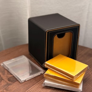 Könnte beinhalten: Eine schwarze quadratische Box mit goldener Innenausstattung und weißen Nähten, neben einem Stapel gelber Karten und transparenten Kunststoffhüllen. Die Artikel befinden sich auf einer dunklen Holzoberfläche.