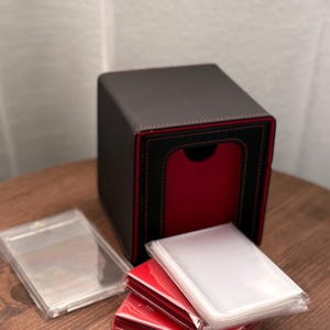 Könnte beinhalten: Eine schwarze und rote Kartenaufbewahrungsbox mit rotem Innenraum und schwarzem Äußeren. Mehrere Stapel Kartenhüllen sind sichtbar, darunter transparente und rote Hüllen. Die Box steht auf einer Holzoberfläche.