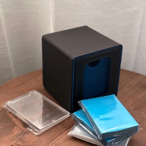 May include: A black and blue card storage box with a blue interior, a clear plastic card holder, and a stack of blue card sleeves. The box is square and sits on a wooden surface.