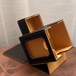May include: A black and tan leatherette desk organizer with a unique, multi-compartment design. The organizer features a rectangular base and a cube-shaped top with open compartments and a window. The interior is tan, and the exterior is black with white stitching.