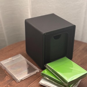 Puede incluir: Una caja de almacenamiento de tarjetas negra, en forma de cubo, con un panel frontal empotrado. Los protectores de tarjetas de plástico transparente y las pilas de fundas verdes y transparentes están junto a la caja sobre una superficie de madera.