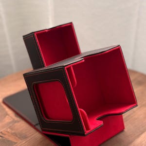 Puede incluir: Caja de almacenamiento de tarjetas negra y roja con tapa con bisagras. La caja tiene un exterior negro, forro interior rojo y costuras. La caja está abierta, revelando los compartimentos interiores. La caja está sobre una superficie de madera.