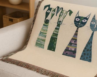 Puede incluir: Manta crema con ilustraciones caprichosas de perros y gatos. Los perros son verdes y los gatos azules, con cuerpos estampados y cuellos largos. La manta tiene un borde con flecos y está sobre un sofá beige.
