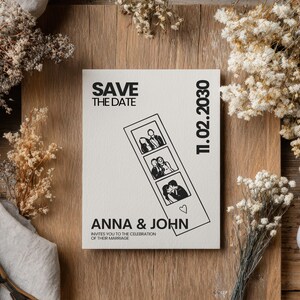 A cream-colored wedding invitation with the text "SAVE THE DATE" and "ANNA & JOHN" in black. The invitation features a photo strip with three images and the date "11.02.2030". The invitation is on a wooden surface with dried flowers.