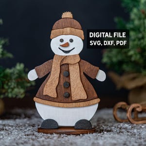Op de afbeelding: Een houten sneeuwpopfiguur met een bruine hoed, sjaal en jas, en een wit lichaam. De sneeuwpop heeft een wortelneus en een lachend gezicht. De tekst "DIGITAL FILE SVG, DXF, PDF" is zichtbaar. Het beeldje staat op een houten voet.
