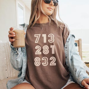 May include: A brown t-shirt with the numbers 713, 281, and 832 printed in a light beige color. The shirt is worn with a denim jacket and sunglasses. A clear glass with a cold beverage is held in the hand.