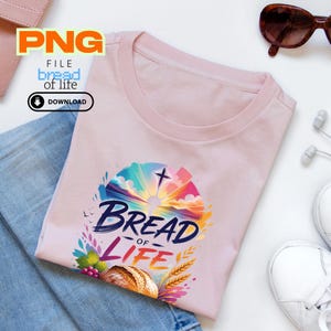 Puede incluir: Camiseta rosa claro con un gráfico colorido que dice "Bread of Life". El gráfico incluye una cruz, un amanecer y una hogaza de pan. La camiseta se combina con vaqueros azules, zapatillas blancas y gafas de sol.