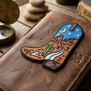 May include: A brown leather journal with a desert-themed patch of a cowboy boot, a compass, and a vintage book titled "Desolation Trails" on a wooden surface. The patch features a cactus, desert landscape, and a night sky.