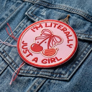 May include: A round, pink embroidered patch with the text "I'M LITERALLY JUST A GIRL" in red lettering. The patch features a cherry design with a red bow and cherries. The patch is sewn onto a blue denim jacket.