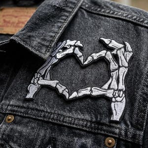 May include: A black denim jacket with a heart-shaped patch made of skeleton hands. The patch is white and gray, and the jacket has a classic collar and button closure. The Levi's tag is visible.