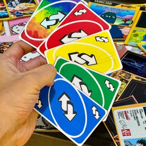 May include: A hand holding a fan of colorful Uno reverse cards. The cards are red, yellow, green, blue, and rainbow-colored, each with a white arrow symbol. Other cards with various designs are visible in the background.