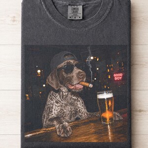 May include: A charcoal gray crew neck t-shirt featuring a graphic print of a spotted dog wearing a black baseball cap and sunglasses, holding a cigar in its mouth while resting its paws on a bar counter next to a glass of beer with a neon sign.