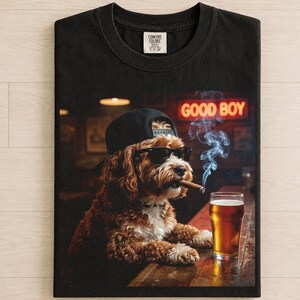 May include: A black t-shirt featuring a graphic of a brown dog wearing a black baseball cap and sunglasses, holding a cigar in its mouth. The dog sits at a bar next to a glass of beer, with a neon sign in the background that reads GOOD BOY.