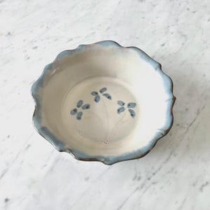 May include: A ceramic bowl with a scalloped edge and a light blue rim. The bowl has a cream-colored interior with three blue floral designs. The bowl's rim is a darker shade of blue, and the overall design is simple and elegant.