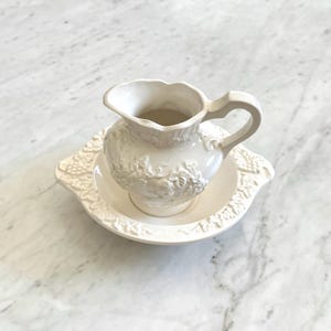 May include: A white ceramic pitcher and basin set. The pitcher has a curved handle and a detailed floral design. The basin has a matching floral design around the rim. The set is placed on a marble surface.