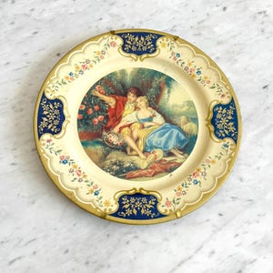May include: Round decorative metal tray with a cream-colored background and a central image of a couple in a garden. The tray has a scalloped edge with floral and blue accents. The central image depicts a man and woman in a garden setting.