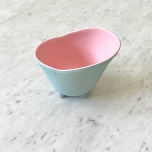 May include: A small, light blue ceramic bowl with a pink interior. The bowl has a textured exterior and rests on three small feet. The interior is a smooth, solid pink. The bowl's shape is reminiscent of a small bathtub.