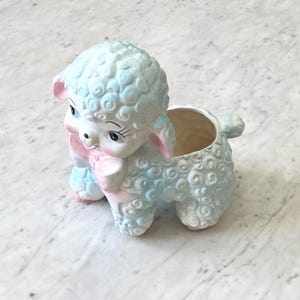 May include: A ceramic planter shaped like a lamb with curly wool texture. The lamb is colored in pale blue and white with pink accents on the ears and a pink bow around its neck. It features large black eyes with long eyelashes.