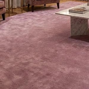Puede incluir: Una alfombra grande y afelpada de color malva cubre el suelo de una sala de estar. La textura de la alfombra es visible y parece suave. Una mesa de centro de m&aacute;rmol y un sof&aacute; malva son parcialmente visibles en el fondo, sugiriendo un interior acogedor y elegante.
