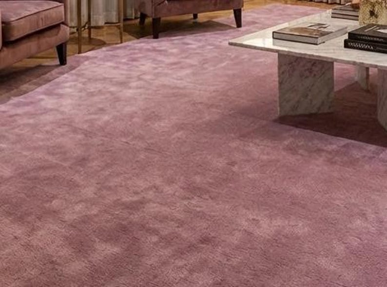 Puede incluir: Una alfombra grande y afelpada de color malva cubre el suelo de una sala de estar. La textura de la alfombra es visible y parece suave. Una mesa de centro de m&aacute;rmol y un sof&aacute; malva son parcialmente visibles en el fondo, sugiriendo un interior acogedor y elegante.