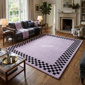 May include: A large, rectangular lavender rug with a black and white checkered border. The rug is placed on a hardwood floor in a living room setting. The text "TORETO RUGS" is visible on the rug.