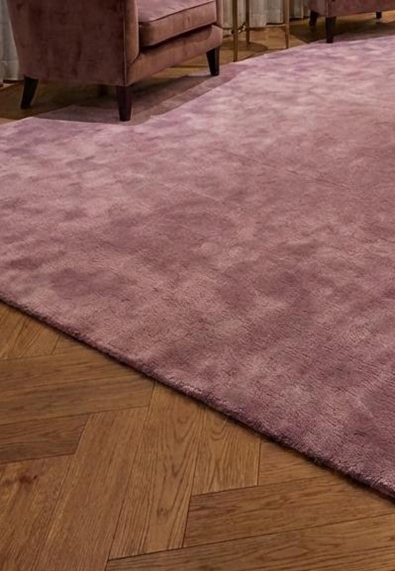 Puede incluir: Una alfombra grande, de color malva, con una superficie suave y texturizada. La alfombra est&aacute; colocada sobre un suelo de madera con un patr&oacute;n de espiga. La alfombra est&aacute; en una habitaci&oacute;n con muebles.