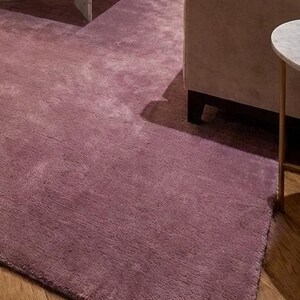 Puede incluir: Primer plano de una alfombra suave de color malva. Se aprecia la textura de la alfombra, con una secci&oacute;n de un sof&aacute; y una peque&ntilde;a mesa auxiliar con tapa de m&aacute;rmol en el fondo. La alfombra est&aacute; sobre un suelo de madera.