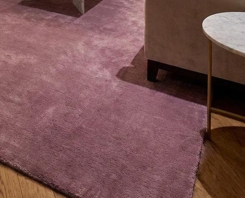 Puede incluir: Primer plano de una alfombra suave de color malva. Se aprecia la textura de la alfombra, con una secci&oacute;n de un sof&aacute; y una peque&ntilde;a mesa auxiliar con tapa de m&aacute;rmol en el fondo. La alfombra est&aacute; sobre un suelo de madera.