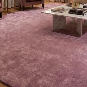 Puede incluir: Una gran alfombra de color malva domina la habitaci&oacute;n. La alfombra es rectangular y cubre la mayor parte del espacio del suelo. La textura parece suave y acogedora. La alfombra est&aacute; rodeada de muebles, incluyendo sillas y una mesa de centro.