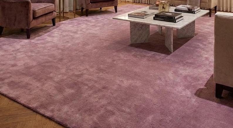 Puede incluir: Una gran alfombra de color malva domina la habitaci&oacute;n. La alfombra es rectangular y cubre la mayor parte del espacio del suelo. La textura parece suave y acogedora. La alfombra est&aacute; rodeada de muebles, incluyendo sillas y una mesa de centro.