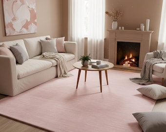 Op de afbeelding: Een gezellige woonkamer met een lichtroze vloerkleed, een witte bank en een open haard. De kamer is ingericht in neutrale tinten met een groot abstract schilderij aan de muur. Een ronde houten salontafel staat in het midden.