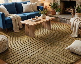 Puede incluir: Una alfombra redonda grande de color verde oliva con un patrón geométrico lineal y texturizado de rayas horizontales y verticales en relieve. La alfombra está colocada sobre un suelo de madera clara en una sala con sofá azul y muebles.