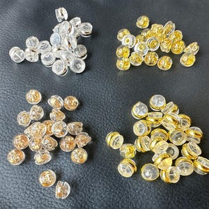 May include: An assortment of earring backs in various styles. The image shows clear, gold, and silver-toned earring backs. The backs are made of clear plastic with metal accents. The earring backs are scattered on a dark surface.