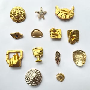 May include: A collection of twelve gold-toned metal buttons and pins featuring various shapes including a lion head, a starfish, a human ear, a nose and lips, a hand holding a handle, a wine glass, a shell, and abstract geometric forms.