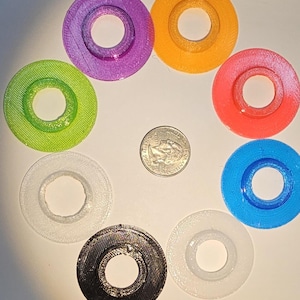 May include: Eight circular, flat, ring-shaped objects in purple, orange, red, blue, white, black, and translucent green and white colors are arranged in a circle around a United States quarter coin for scale, showing their textured surfaces.