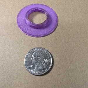 May include: A purple 3D-printed circular washer with a raised center ring, positioned next to a United States quarter coin for scale. The object features a textured surface and a hollow center, showing a diameter slightly larger than the 0.955 inch coin.