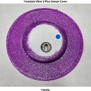 May include: A circular purple Freestyle Libre 3 Plus Sensor Cover with a textured surface. The center features a white circular area with a small metallic component and a single blue dot. The text Freestyle Libre 3 Plus Sensor Cover is visible.