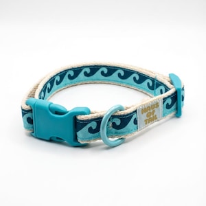 Peut inclure: Collier pour chien bleu et crème avec un motif de vagues. Il est équipé d'une boucle en plastique turquoise et d'un anneau métallique pour l'attache d'une laisse. La marque "Haus of Tail" est imprimée sur une petite étiquette.