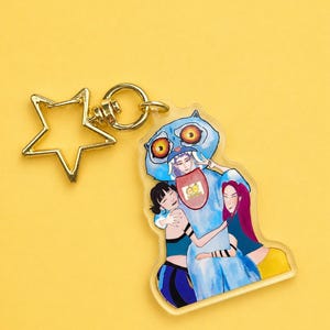 Peut inclure: Un porte-clés en acrylique transparent représentant un personnage de dessin animé bleu étreignant deux personnages. Le porte-clés a un charm en forme d'étoile dorée et un anneau doré. Le fond est jaune uni.