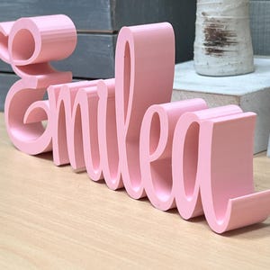 May include: A pink, cursive-style wooden sign that spells out the name "Emilea." The letters are thick and stand upright on a light-colored wooden surface. The sign is a decorative item, possibly for a child's room or a personalized gift.